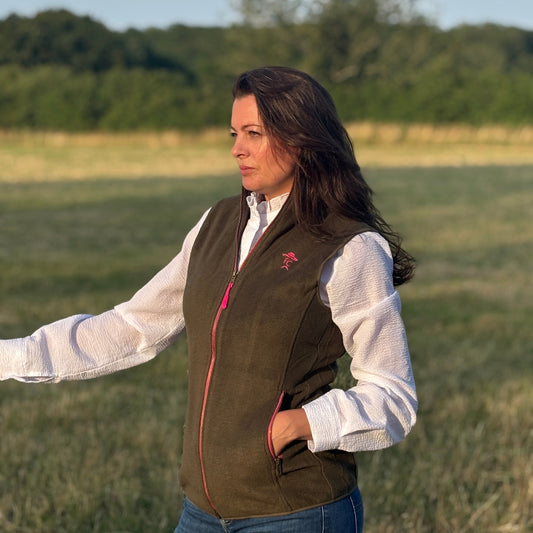 Woman wearing a brown vest over a white shirt in an open field with trees in the background