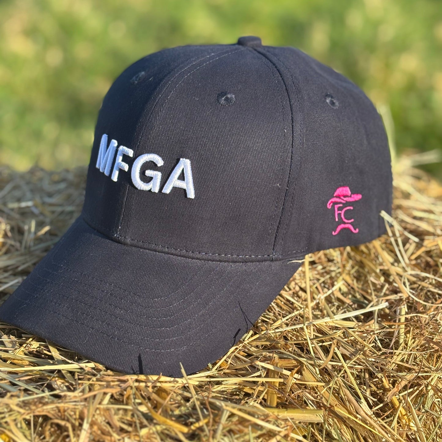 Black cap with 'MFGA' and pink logo on a hay bale