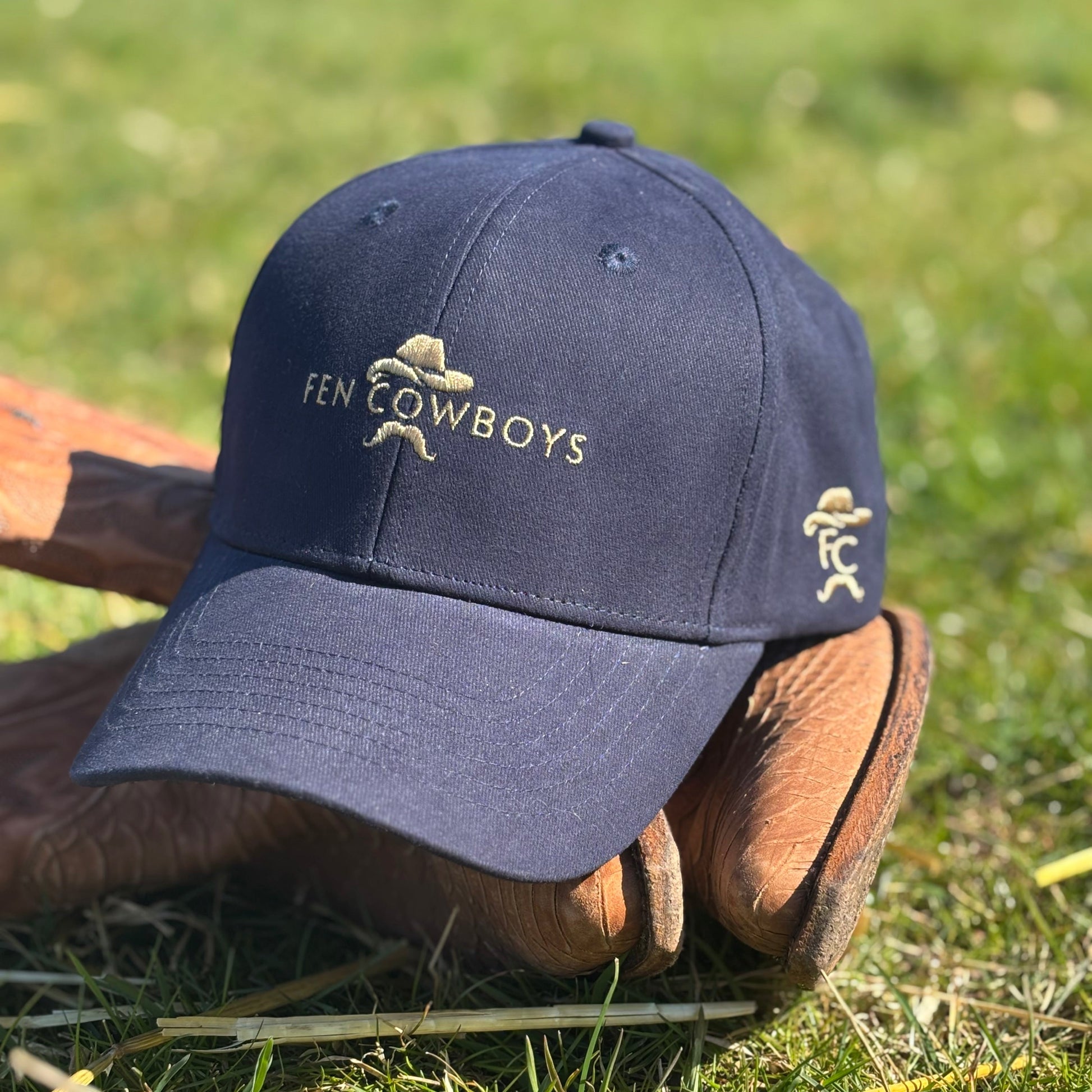 Navy blue cap with 'Fen Cowboys' logo on a wooden boot in a grassy field