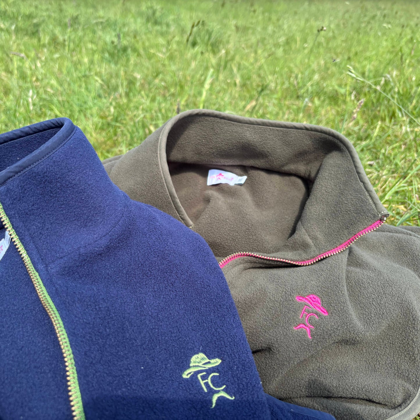 Two fleece jackets, one blue and one gray, with visible branding on a grassy background.
