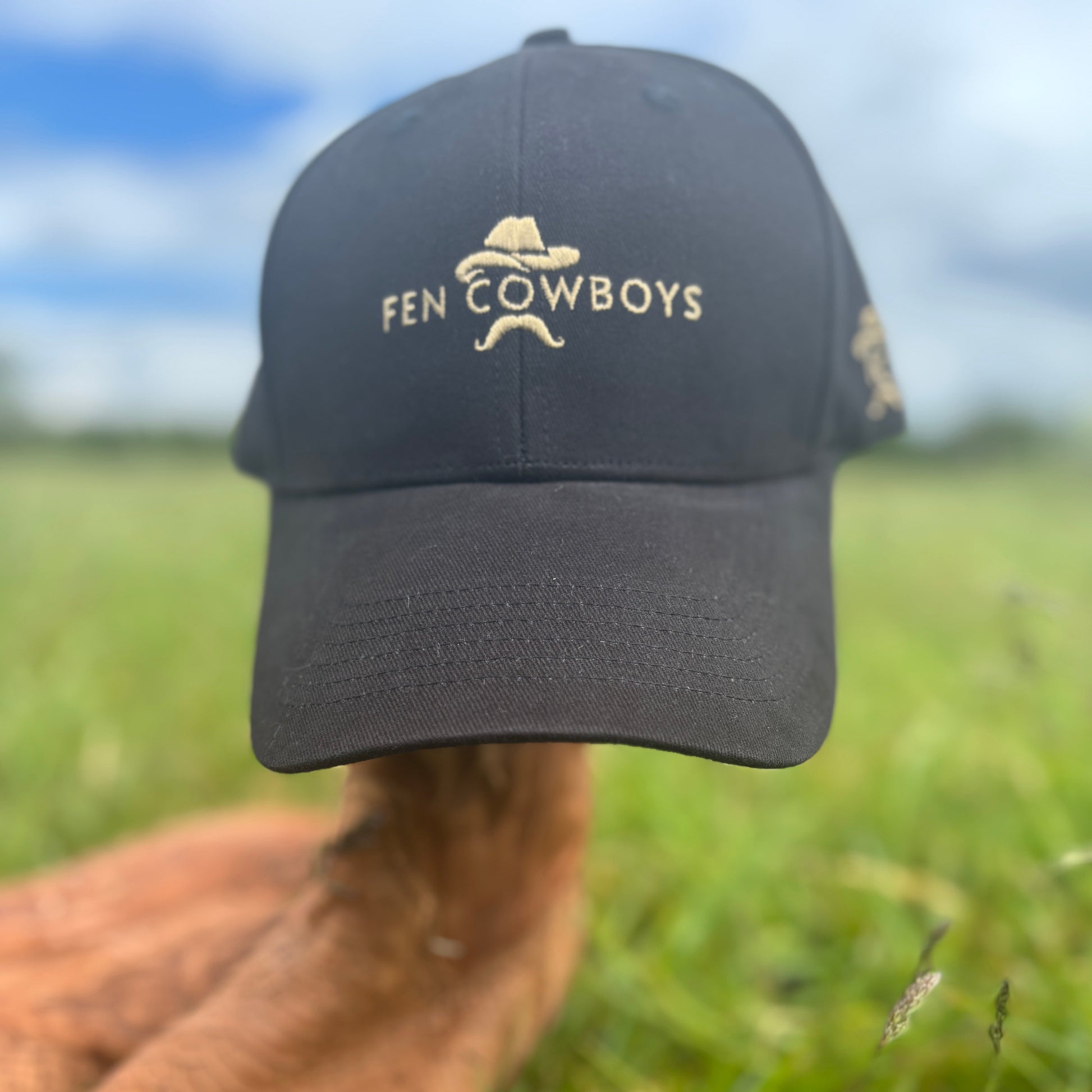 Black cap with 'Fen Cowboys' logo held by a hand in a grassy field