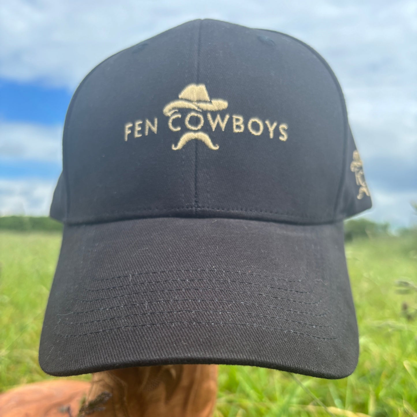 Black cap with 'Fen Cowboys' logo held by a person in a grassy outdoor setting