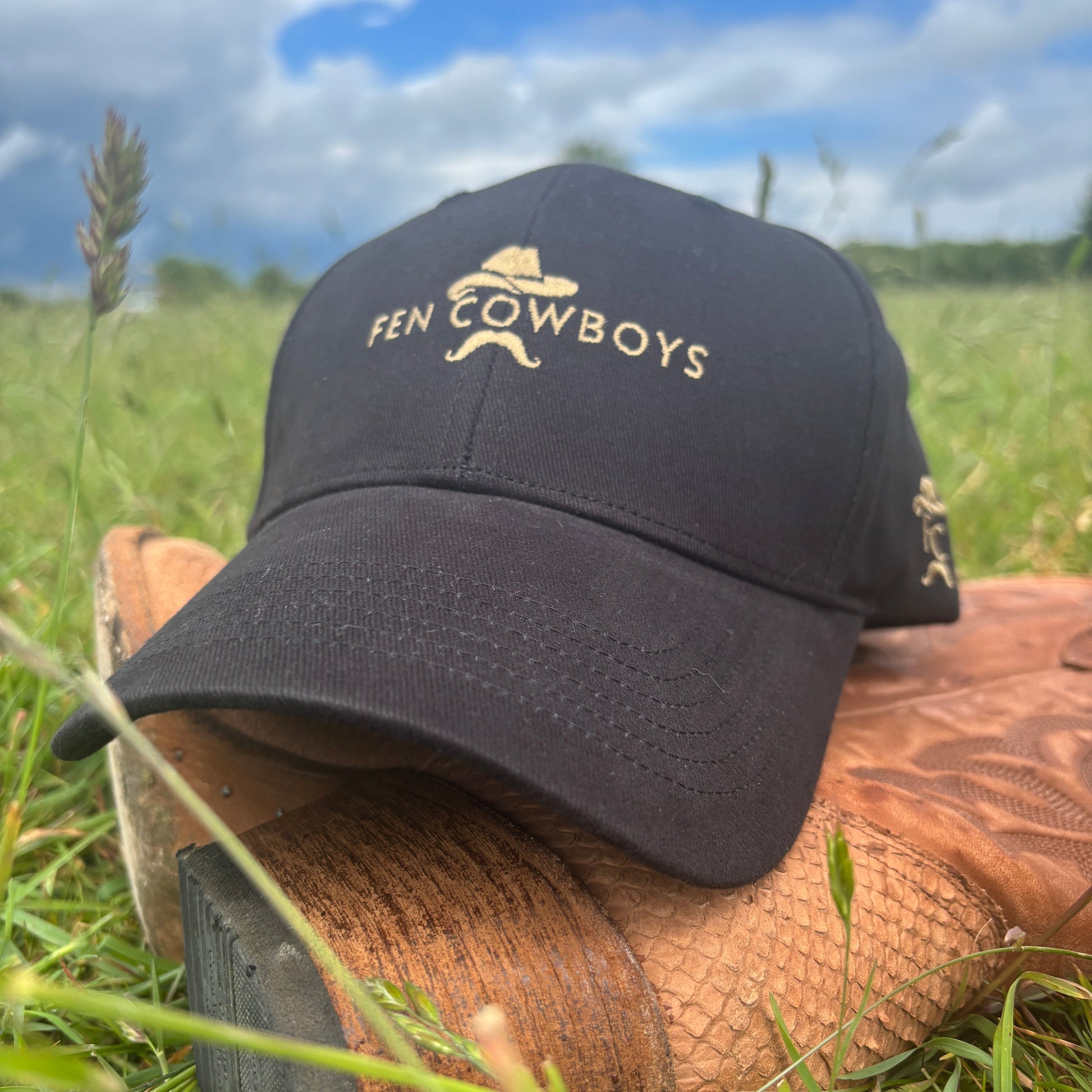 Black cap with 'Fen Cowboys' logo on a wooden duck call against a grassy background