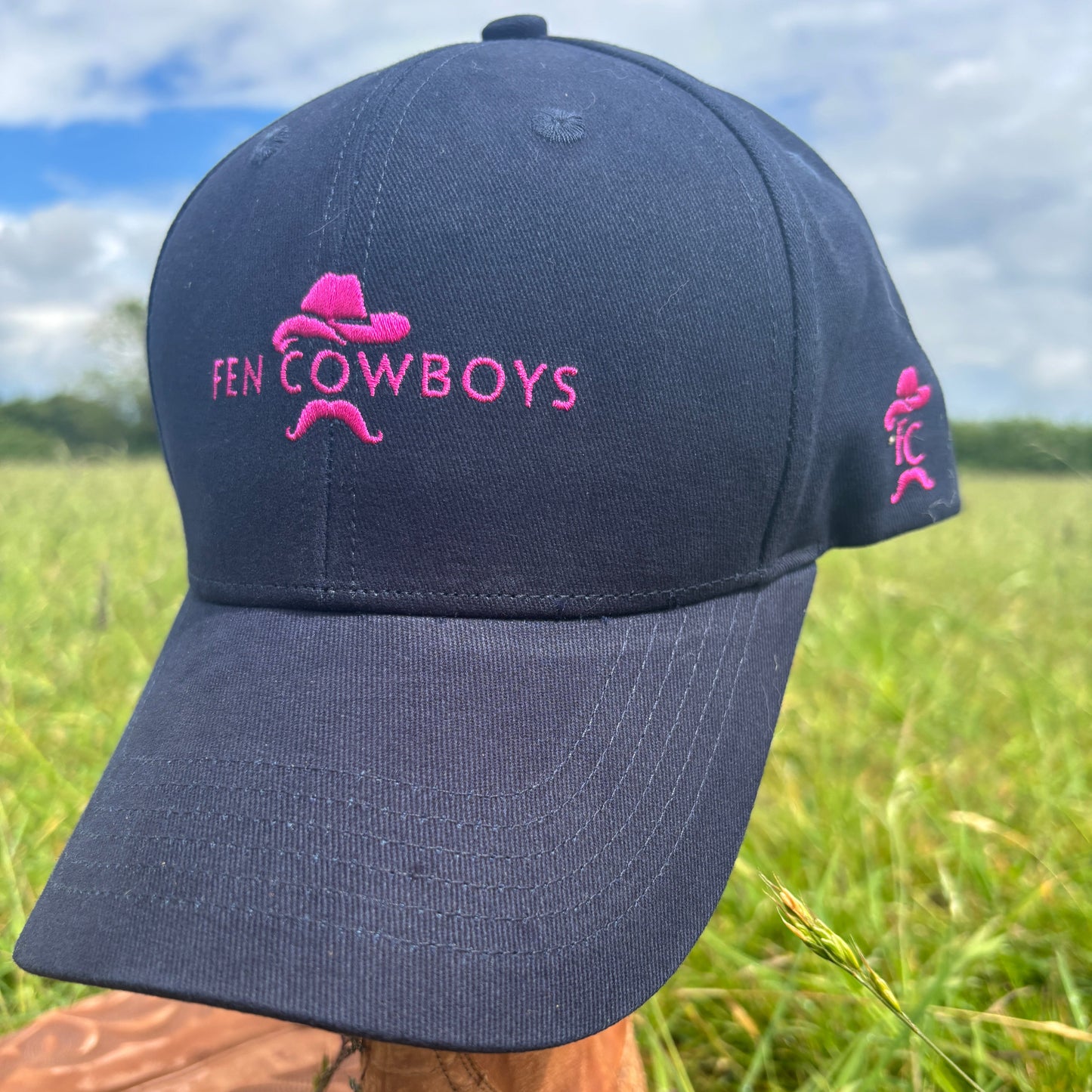Black cap with pink 'Fen Cowboys' logo held by a hand in grass