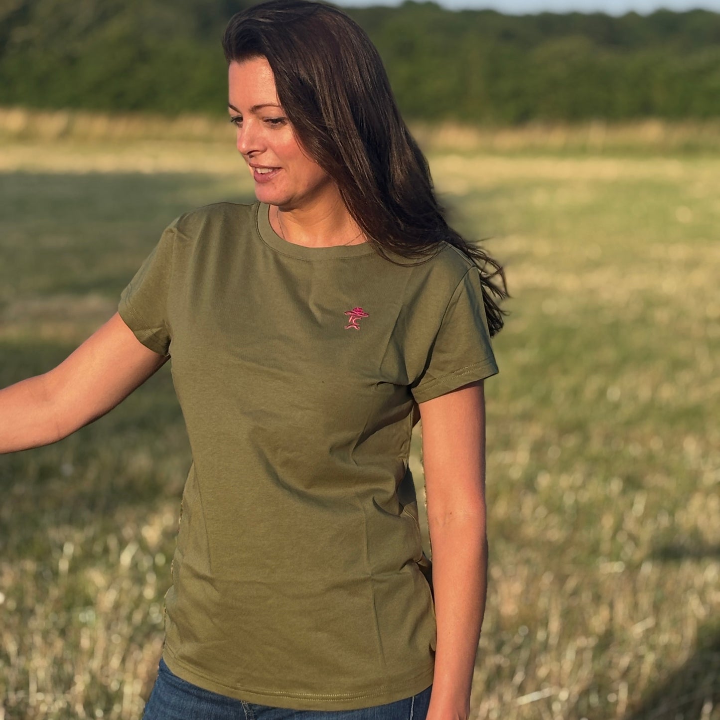 Woman wearing a green t-shirt with a small red logo, standing in a field.