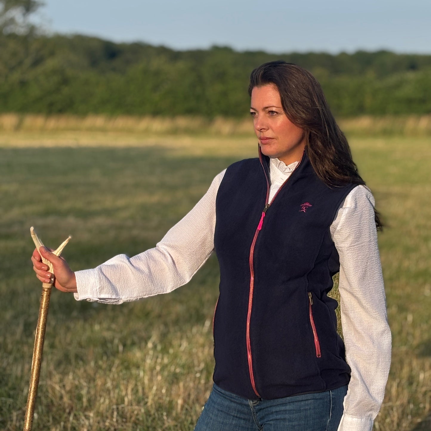 Woman standing in a field holding a stick, wearing a navy vest over a white shirt.