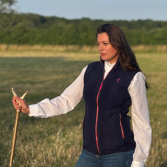 Woman standing in a field holding a stick, wearing a navy vest over a white shirt.