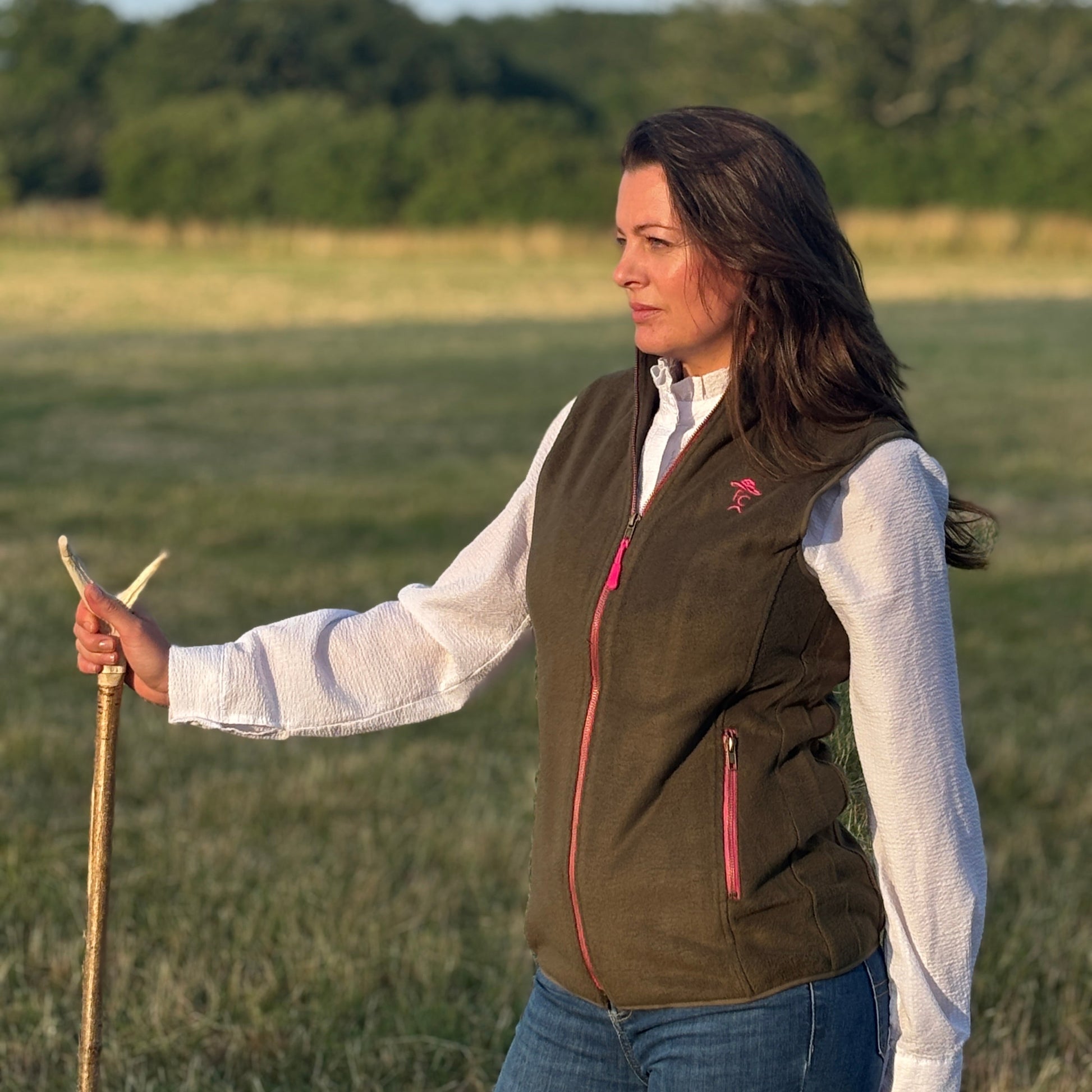 Woman standing in a field wearing a green vest and holding a long stick.