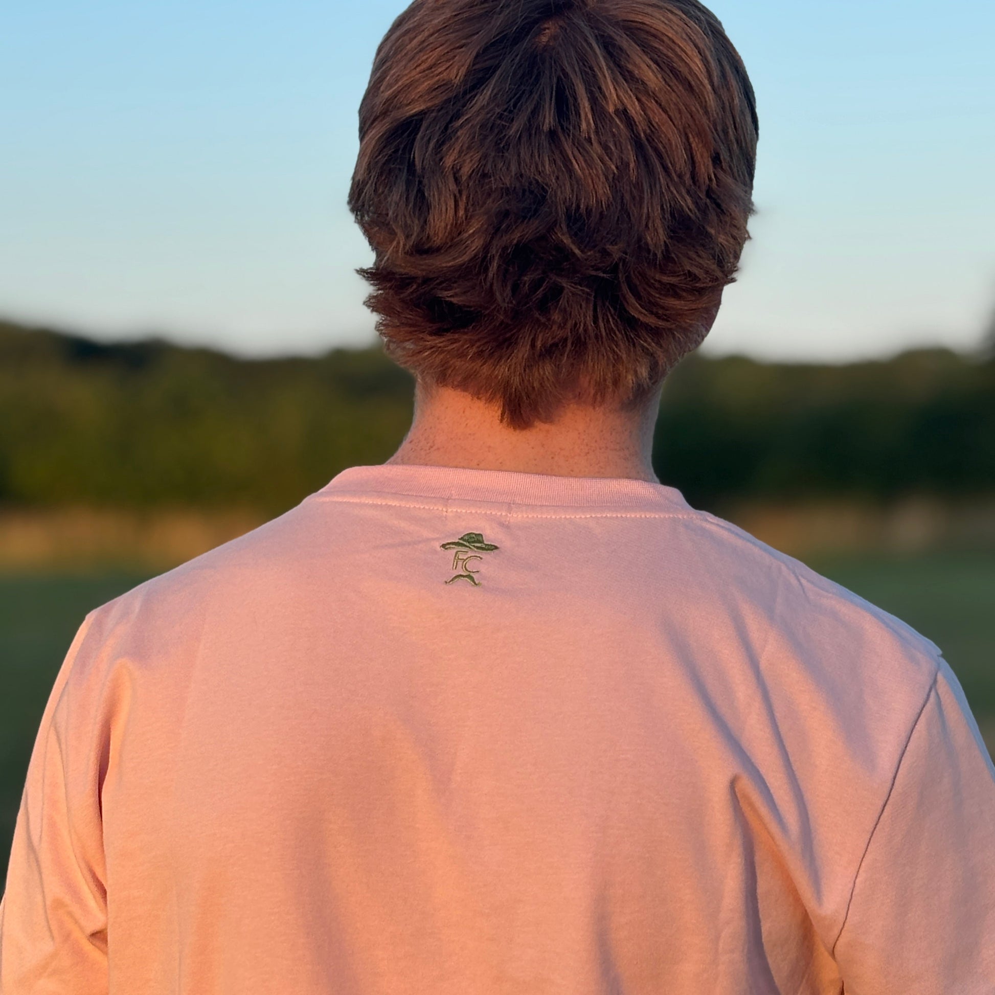 Person wearing a light pink Fen Cowboys t-shirt with a small tree design on the back, standing in a field.