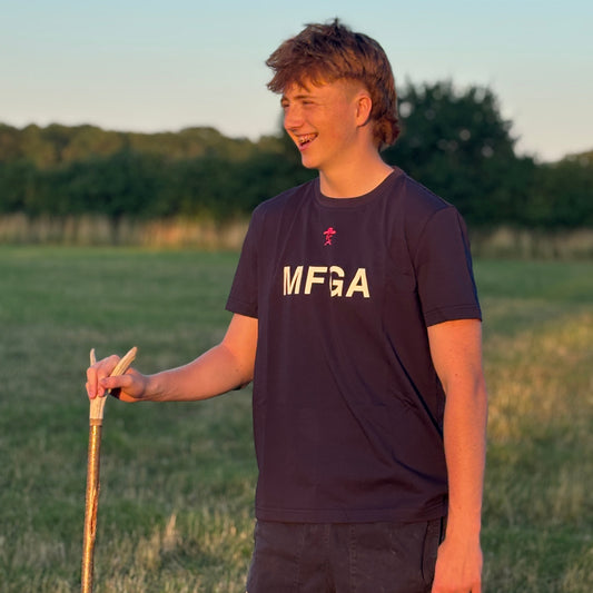 Person wearing a dark blue t-shirt with 'MFGA' logo, holding a walking stick in a field.