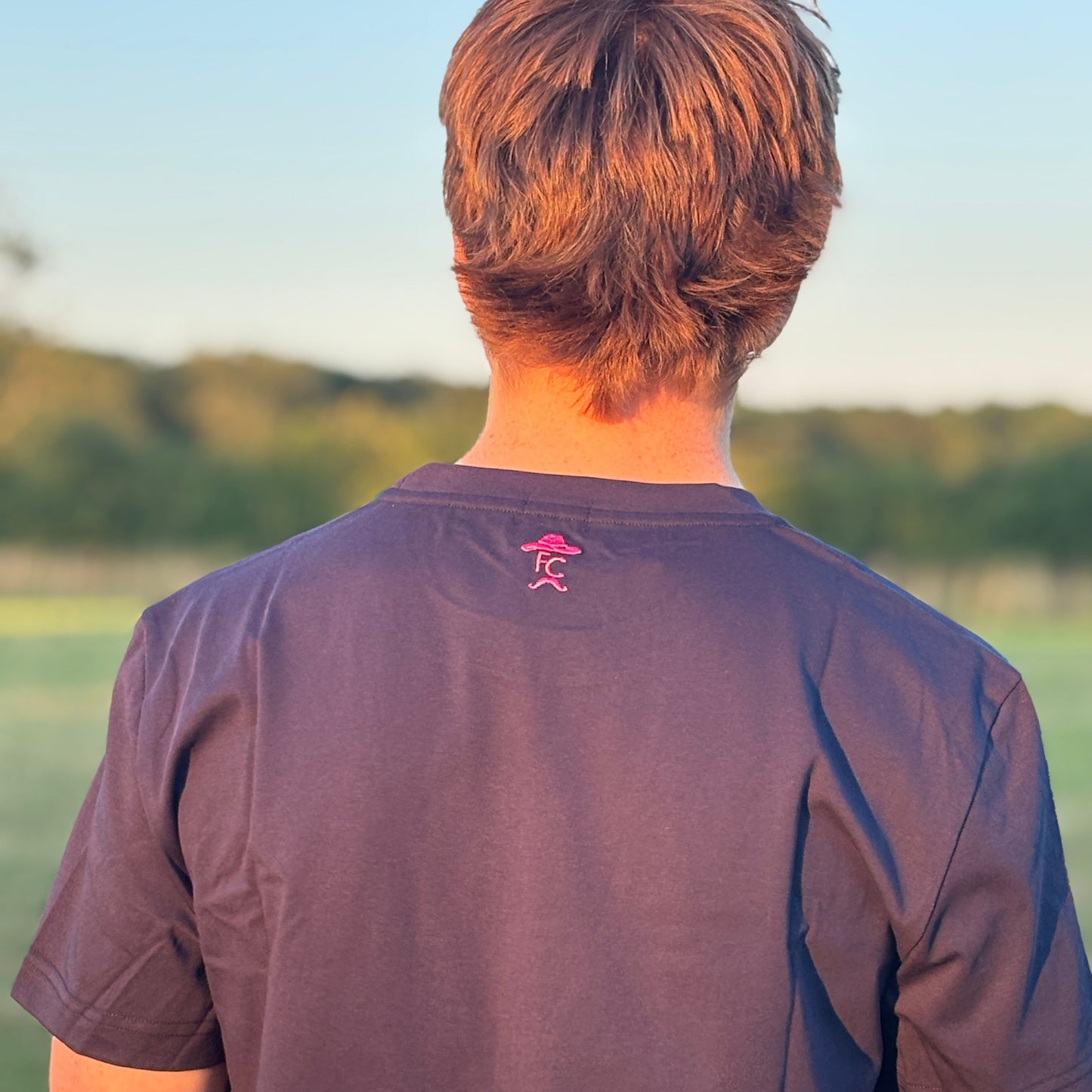 Person wearing a dark blue t-shirt with a pink logo, standing outdoors with a scenic background.