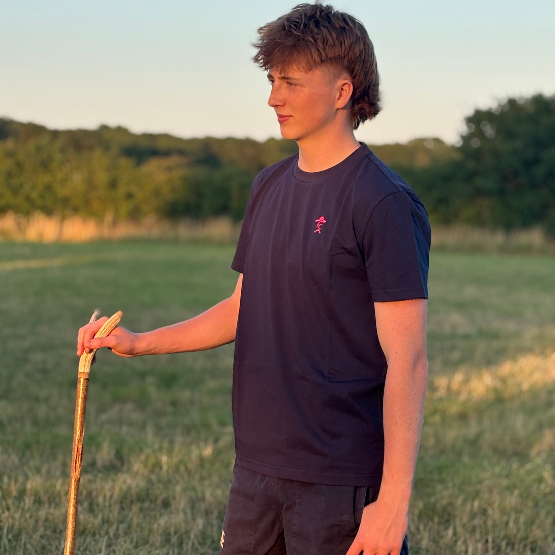 Person standing in a field holding a walking stick, wearing a Fen Cowboys dark t-shirt and shorts.