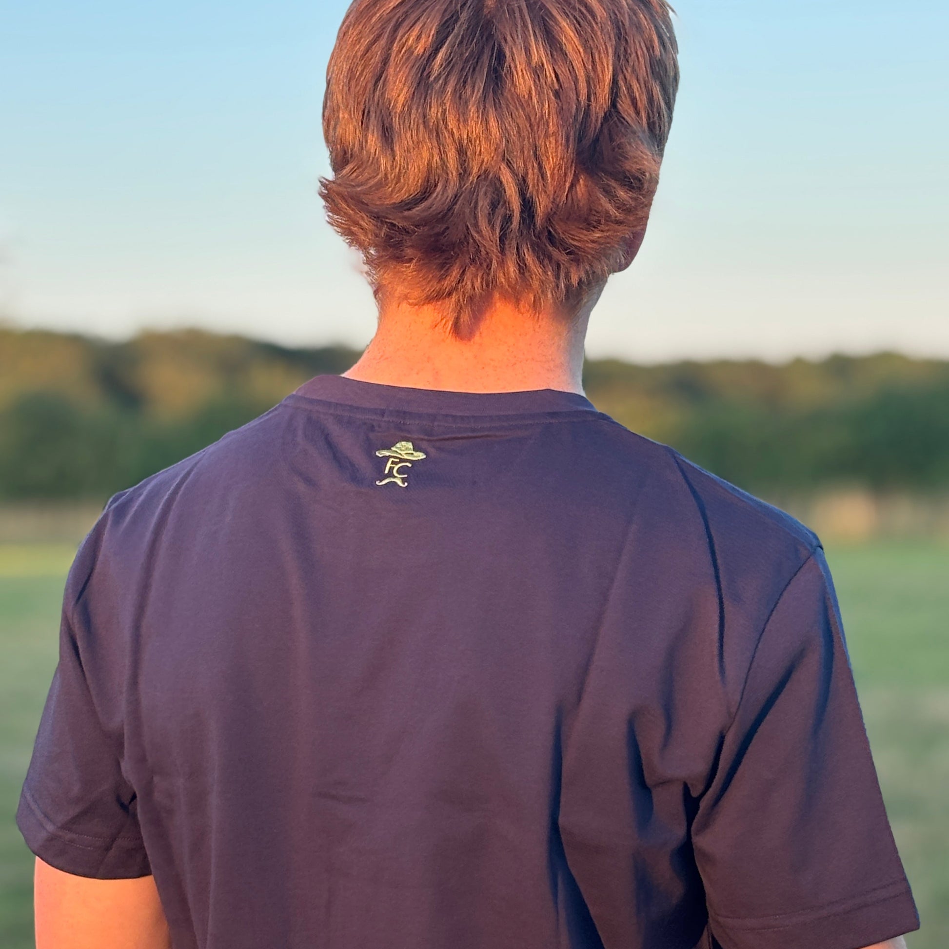 Person wearing a Fen Cowboys dark blue t-shirt with a logo on the back, standing outdoors with a blurred natural background.
