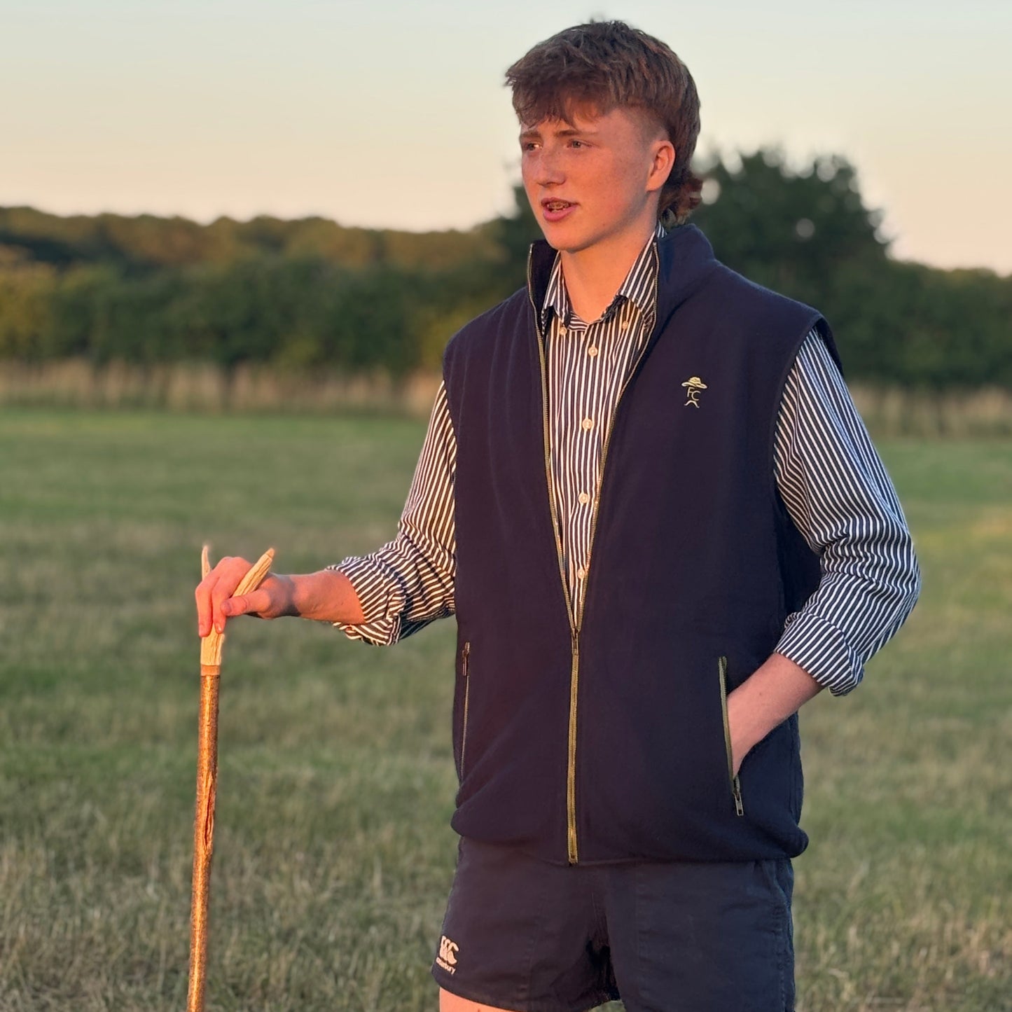 Person standing in a field wearing a Fen Cowboys gilet and shorts, holding a walking stick.