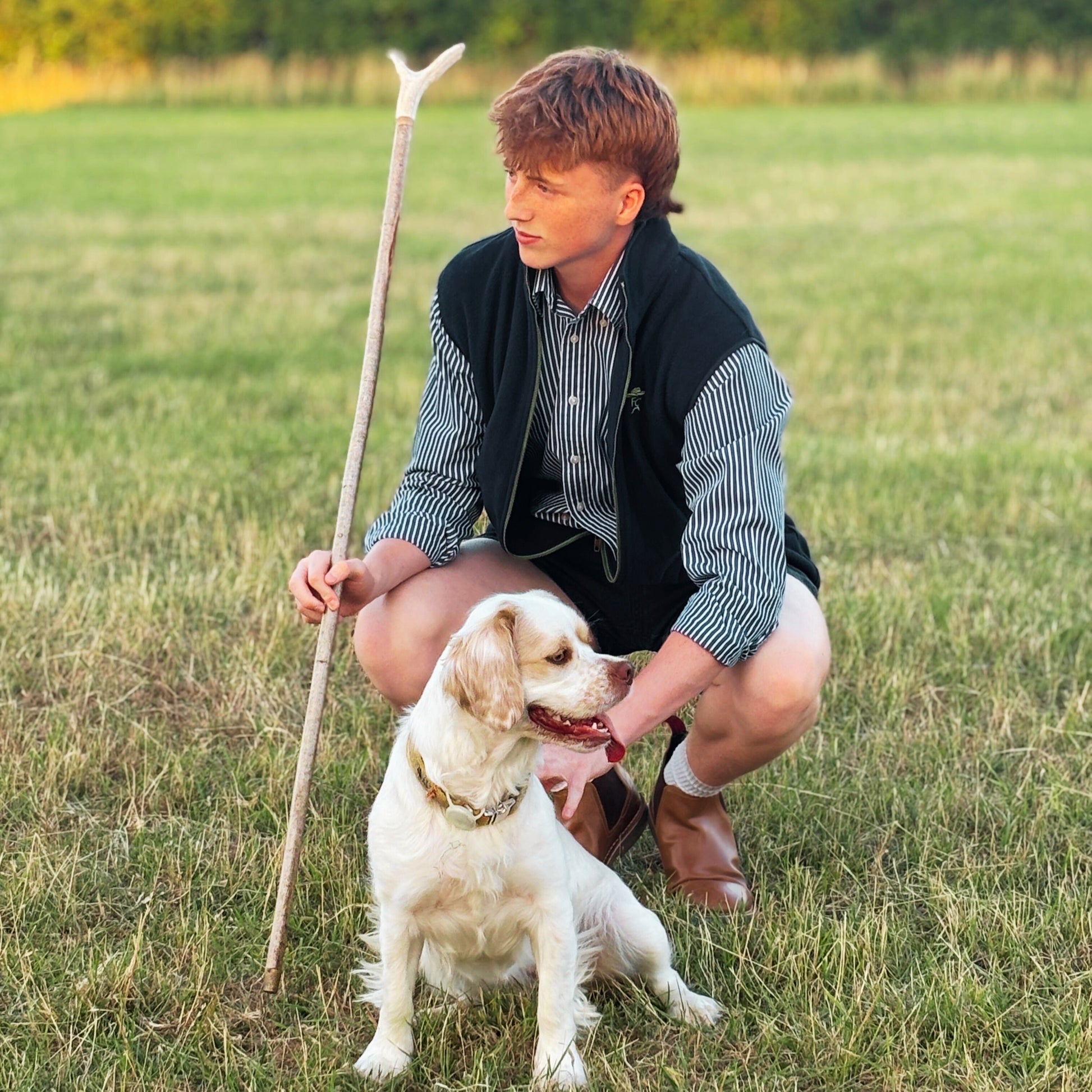 Man crouching with a dog in a field holding a stick wearing Fen Cowboys gilet
